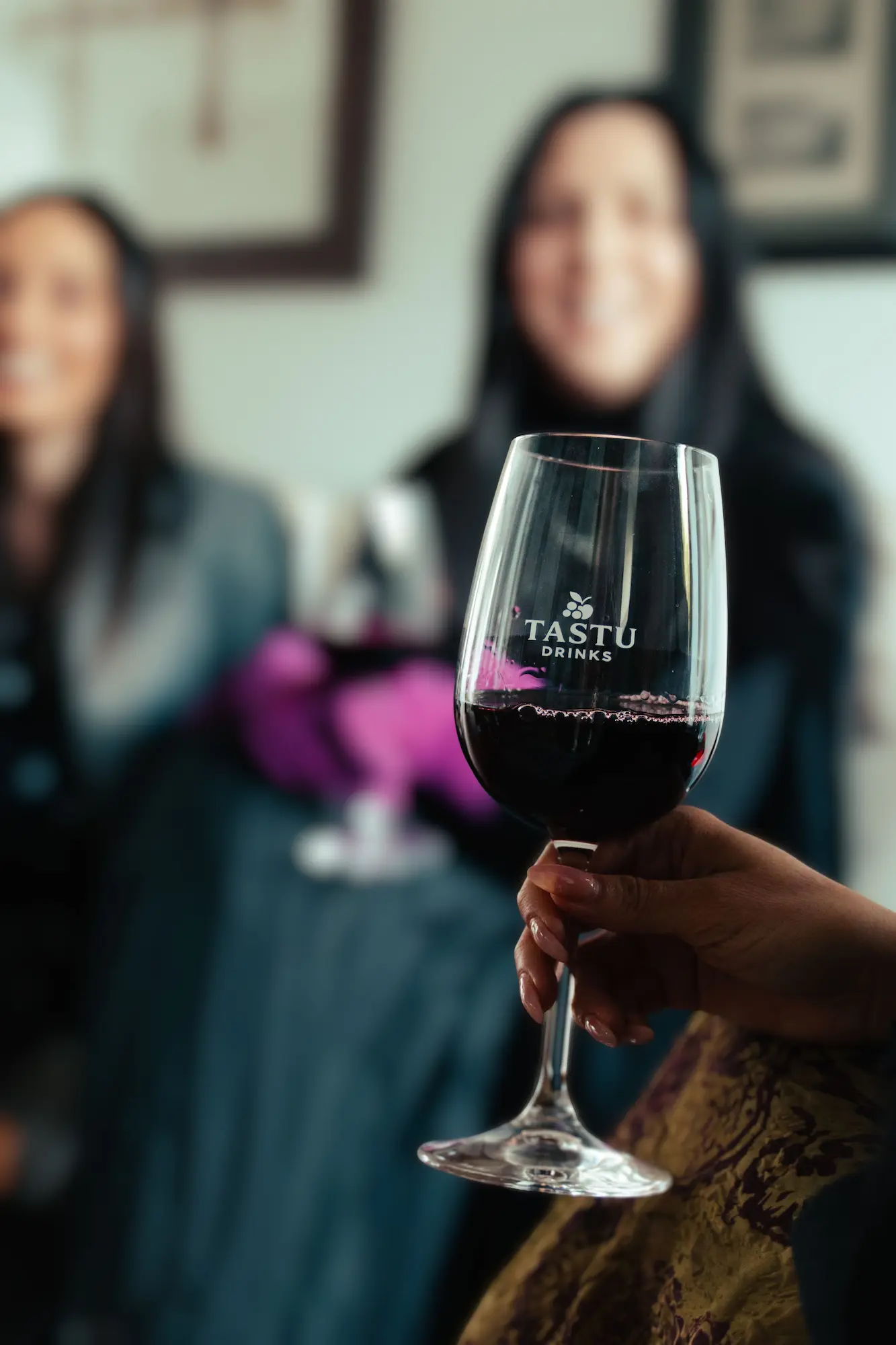 Primer plano de dos copas de vino tinto con el logo de TASTU DRINKS chocando en un brindis. Una mano lleva un guante fucsia.