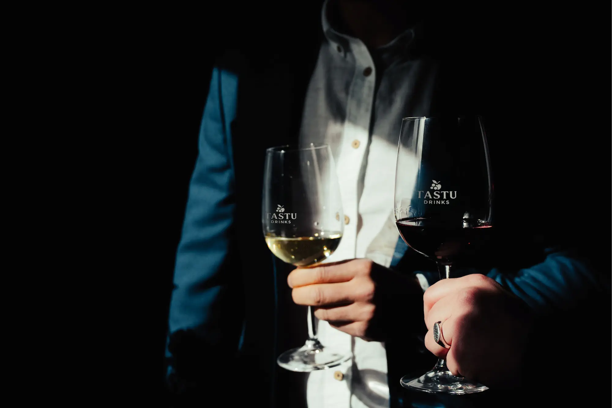 Tastu Drinks - Estaciones de Vino y Sidra Un hombre de traje sosteniendo una copa de vino tinto y otra de vino blanco en un ambiente oscuro.