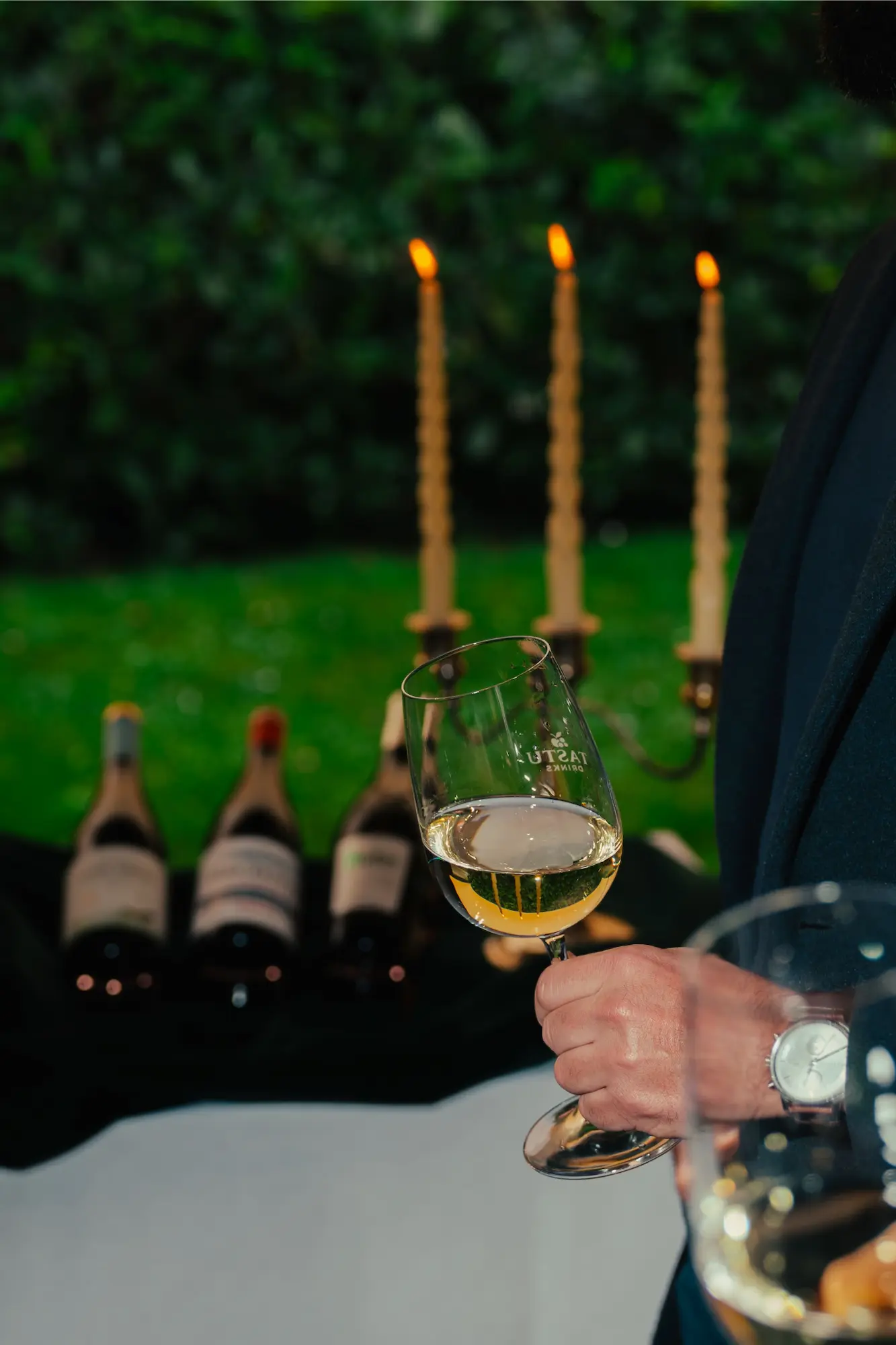 Tastu Drinks - Estaciones de Vino y Sidra Hombre elegante sujetando una copa de vino junto a botellas Tastu y velas en una mesa de madera.