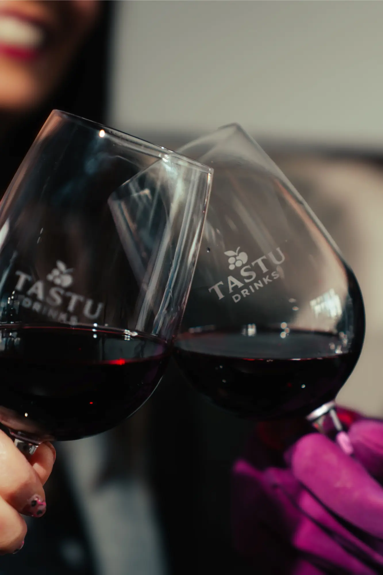 Tastu Drinks - Estaciones de Vino y Sidra Primer plano de una mano con guante rosa sosteniendo una copa de vino tinto frente a un viñedo al atardecer.