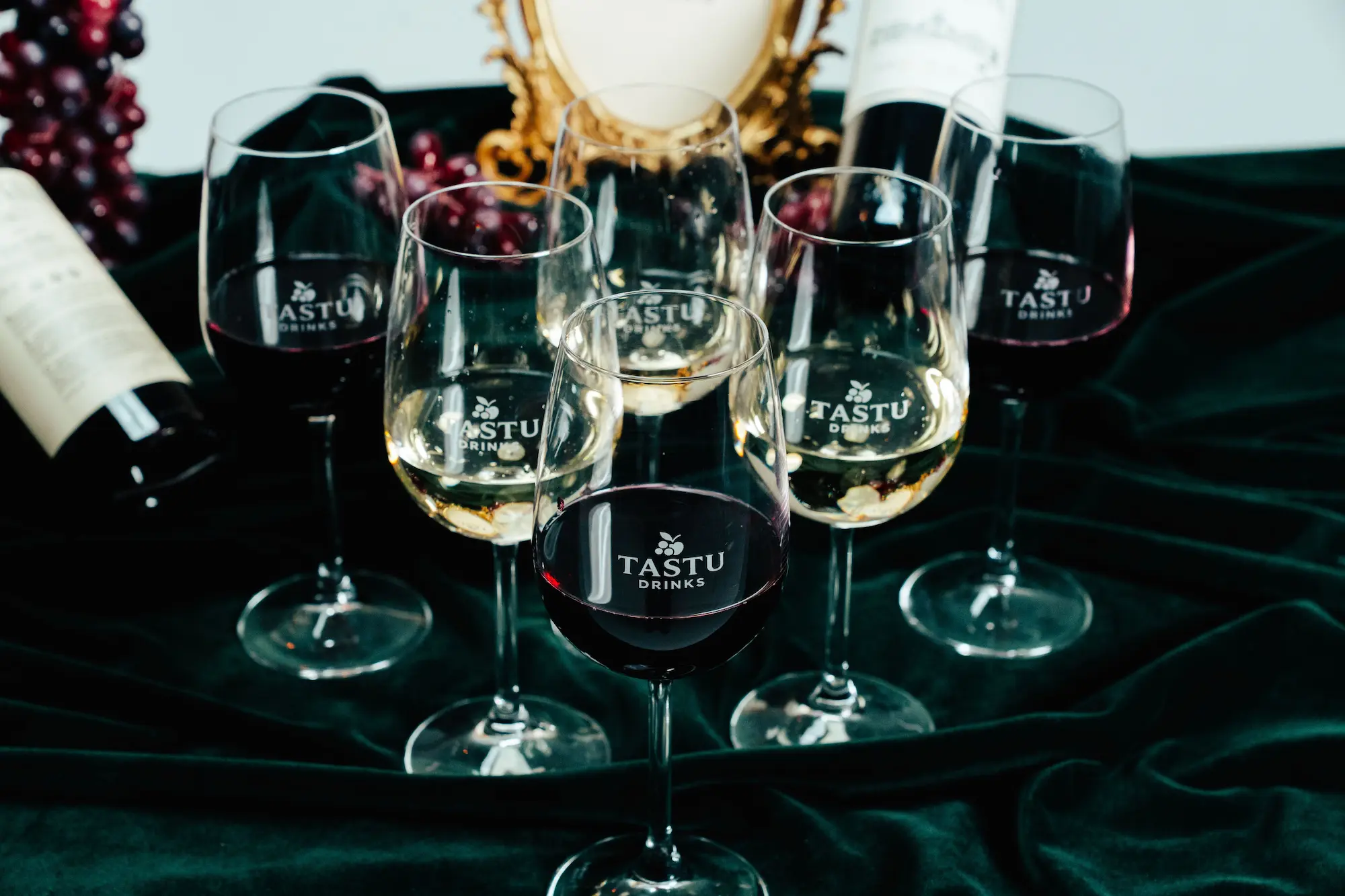 Tastu Drinks - Estaciones de Vino y Sidra Bodegón de Tastu Drink con dos botellas de vino, copas, uvas y una tabla de quesos sobre madera.