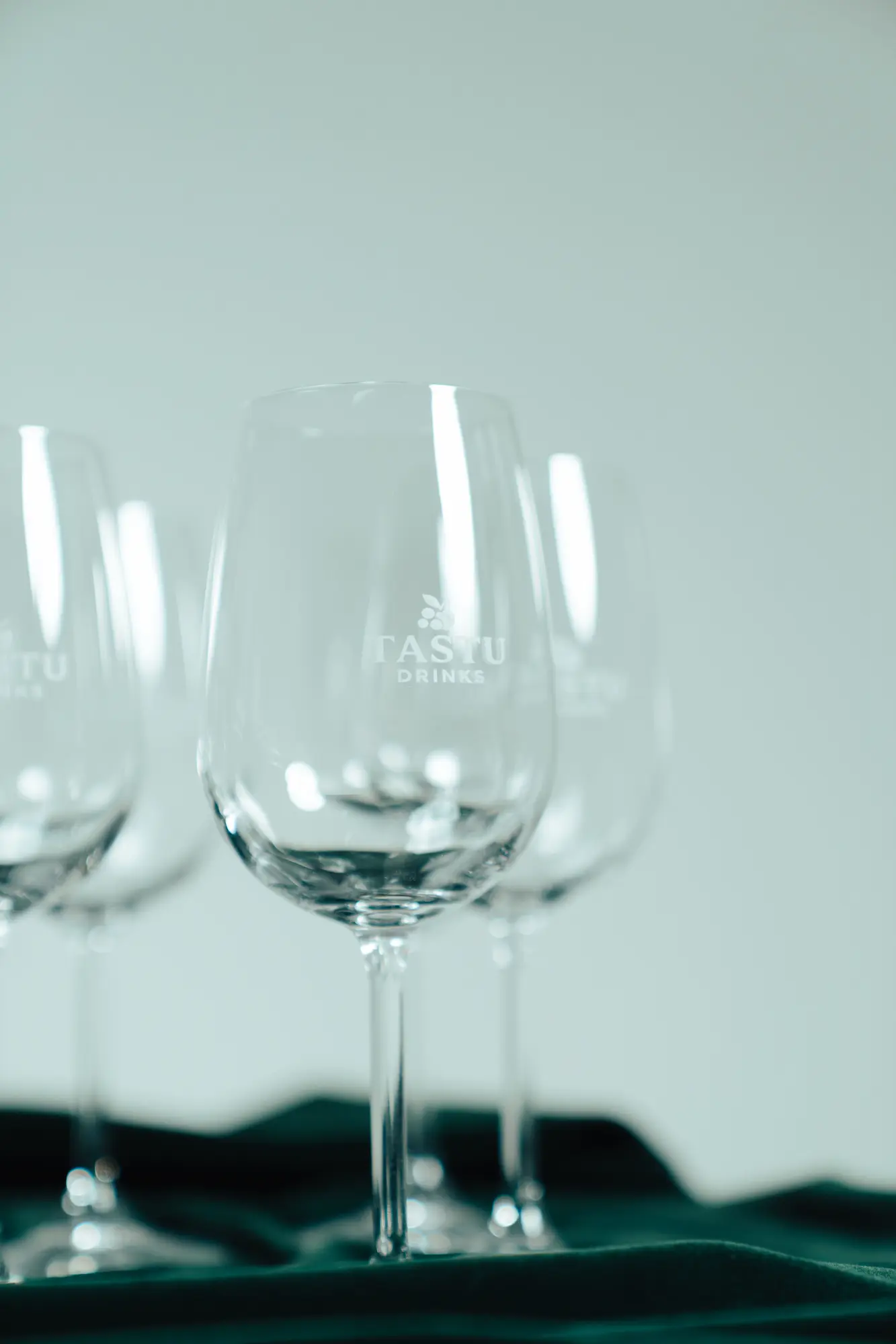 Varias copas de cristal vacías con logo Tastu sobre una mesa de madera y fondo desenfocado.